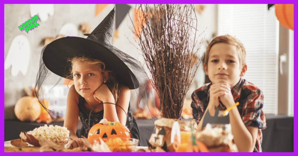 Children dressed for Halloween sitting at a decorated table with pumpkins and popcorn, one wearing a witch hat.