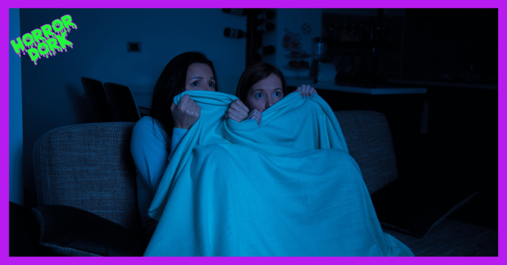 Two people on a couch watching a scary movie at night, hiding under a blanket with wide, frightened eyes; Horror Dork logo in the corner.